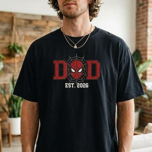 Pu&ograve; includere: T-shirt nera con un design grafico rosso e bianco. La parola "DAD" &egrave; in lettere rosse con una ragnatela e una maschera di Spider-Man al centro. Il testo "EST. 2026" &egrave; sotto.