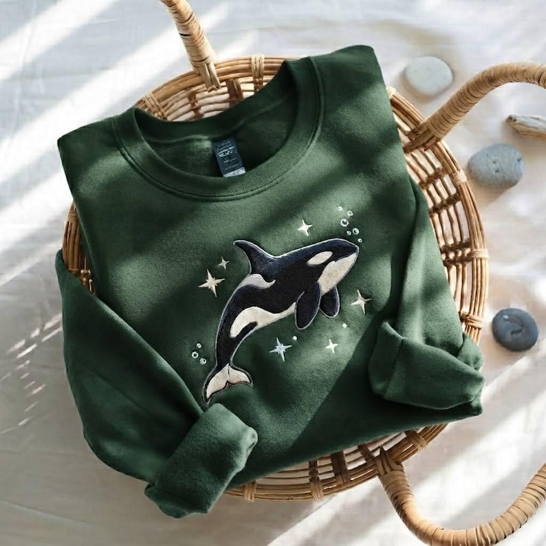 May include: A dark forest green crewneck sweatshirt featuring an embroidered black and white orca whale graphic centered on the chest. The orca is surrounded by small embroidered white stars and circular bubbles, creating a celestial ocean theme.