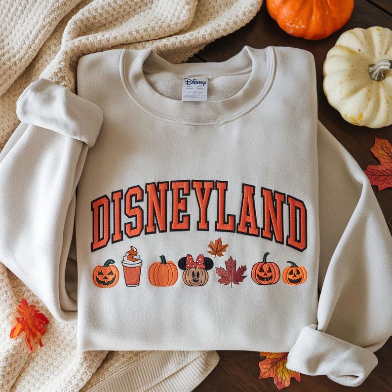 May include: Cream-colored sweatshirt with the word "DISNEYLAND" in orange block letters. Below the text are embroidered pumpkins, a pumpkin spice latte, a Minnie Mouse pumpkin, and fall leaves. The sweatshirt is folded on a cream-colored blanket.