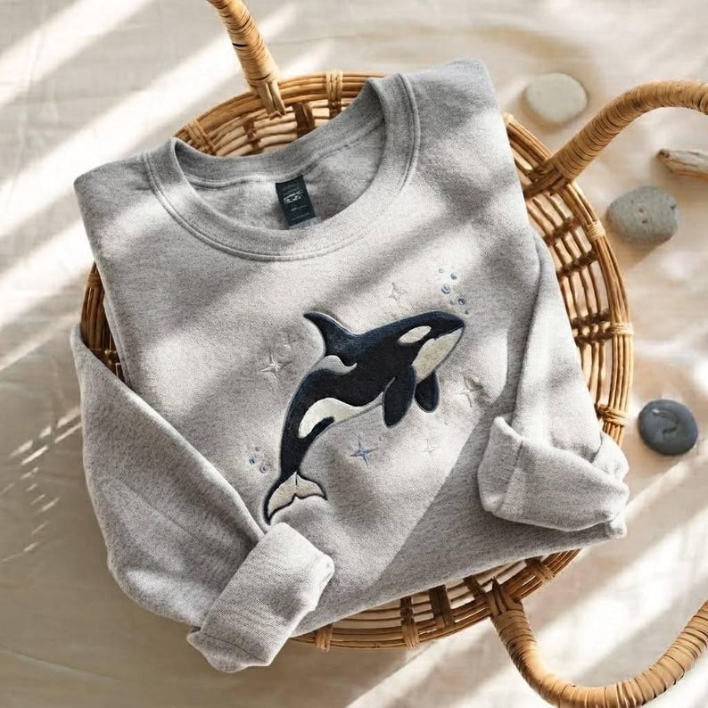 May include: A heather gray crewneck sweatshirt featuring an embroidered black and white orca whale graphic centered on the chest. The design includes small embroidered stars and bubbles surrounding the whale, with long sleeves and ribbed cuffs.