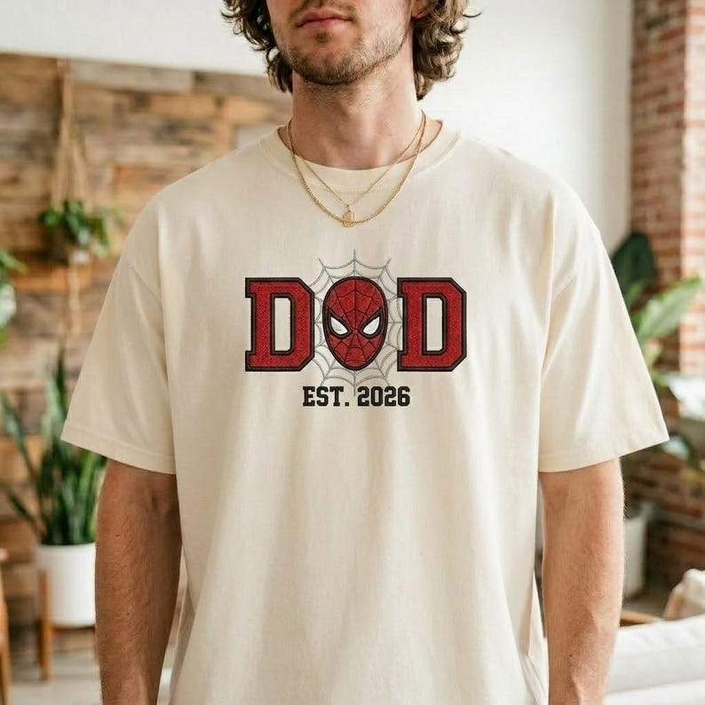 Pu&ograve; includere: T-shirt color crema con la scritta "DAD" in lettere rosse con contorno nero. La "O" &egrave; sostituita da un disegno della maschera di Spider-Man in una ragnatela. Sotto, "EST. 2026".