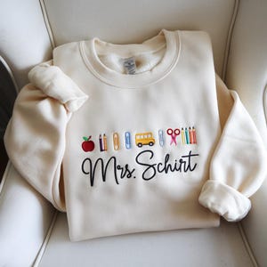 Pode incluir: Camisola creme com gráficos bordados com tema escolar, incluindo uma maçã, lápis, clipes de papel, um autocarro escolar, tesouras e lápis de cor. As palavras "Mrs. Schirt" são bordadas abaixo.
