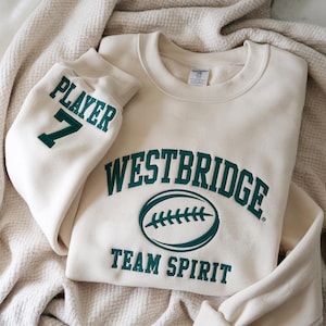 Puede incluir: Sudadera crema con las palabras "WESTBRIDGE TEAM SPIRIT" en letras verde oscuro. La manga izquierda muestra "PLAYER 7" en el mismo verde. El diseño incluye un gráfico de fútbol americano. La sudadera está doblada sobre una manta crema texturizada.