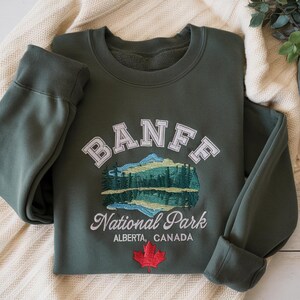 May include: A dark olive green crewneck sweatshirt featuring embroidered text that reads BANFF National Park ALBERTA, CANADA. The design includes a mountain and forest landscape graphic and a small red maple leaf icon below the text.