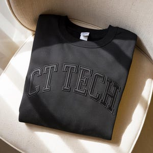 May include: A black sweatshirt featuring the embroidered text "CT TECH" in a collegiate font. The garment is folded on a cream-coloured chair, highlighting the design and fabric texture.