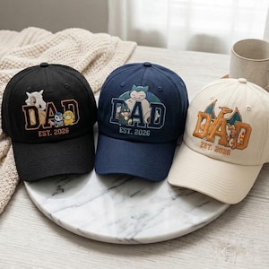 May include: Three baseball caps in black, navy blue, and cream. Each cap features the word "DAD" in an orange embroidered font, with a cartoon character design and the text "EST. 2026". The caps are displayed on a marble-patterned surface.