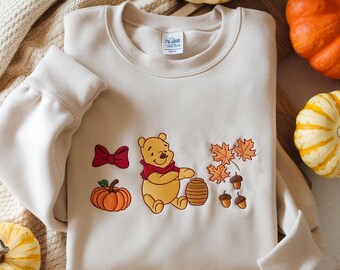 May include: Cream-colored sweatshirt featuring an embroidered Winnie the Pooh design with a red bow, pumpkin, maple leaves, acorns, and a honey pot. The long-sleeved top is surrounded by pumpkins.