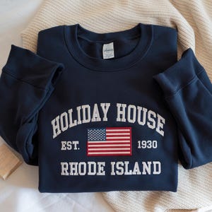 May include: Navy blue sweatshirt with the words "HOLIDAY HOUSE" and "RHODE ISLAND" in white block letters. A small American flag patch is centered below the text, with the year "1930" printed to the right. The sweatshirt is folded.