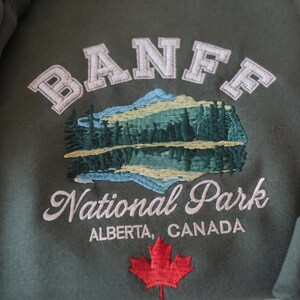 May include: An olive green sweatshirt featuring an embroidered design of Banff National Park. The text reads BANFF, National Park, and ALBERTA, CANADA, accompanied by a landscape of mountains, trees, and a red maple leaf emblem.