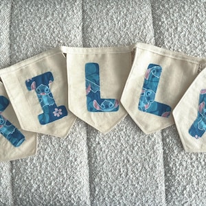 K&ouml;nnte beinhalten: Ein Banner mit der Aufschrift "LILY" mit einzelnen Wimpeln. Jeder Wimpel ist cremefarben mit einem blauen Buchstaben. Die Buchstaben sind mit einem blauen und rosa Cartoon-Charakter-Design verziert. Das Banner wird auf einer strukturierten, hellfarbigen Oberfl&auml;che angezeigt.