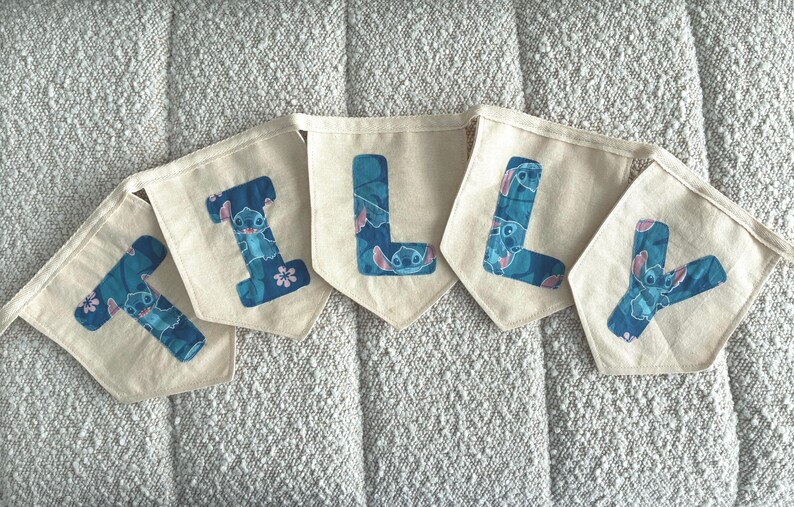 K&ouml;nnte beinhalten: Ein Banner mit der Aufschrift "LILY" mit einzelnen Wimpeln. Jeder Wimpel ist cremefarben mit einem blauen Buchstaben. Die Buchstaben sind mit einem blauen und rosa Cartoon-Charakter-Design verziert. Das Banner wird auf einer strukturierten, hellfarbigen Oberfl&auml;che angezeigt.