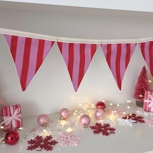 May include: A fabric bunting banner featuring triangular pennants with alternating vertical stripes in shades of red and pink, suspended from a white cord. The banner is displayed horizontally with a series of red and pink holiday decorations.