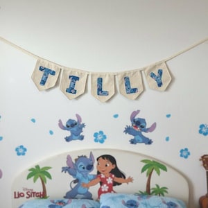 K&ouml;nnte beinhalten: Ein Banner mit der Aufschrift "TILLY" h&auml;ngt &uuml;ber einem Bett mit dem Disney Lilo & Stitch-Thema. Die Buchstaben sind blau auf beigem Hintergrund. Das Kopfteil zeigt Lilo und Stitch, Palmen und die Worte "Lilo & Stitch".