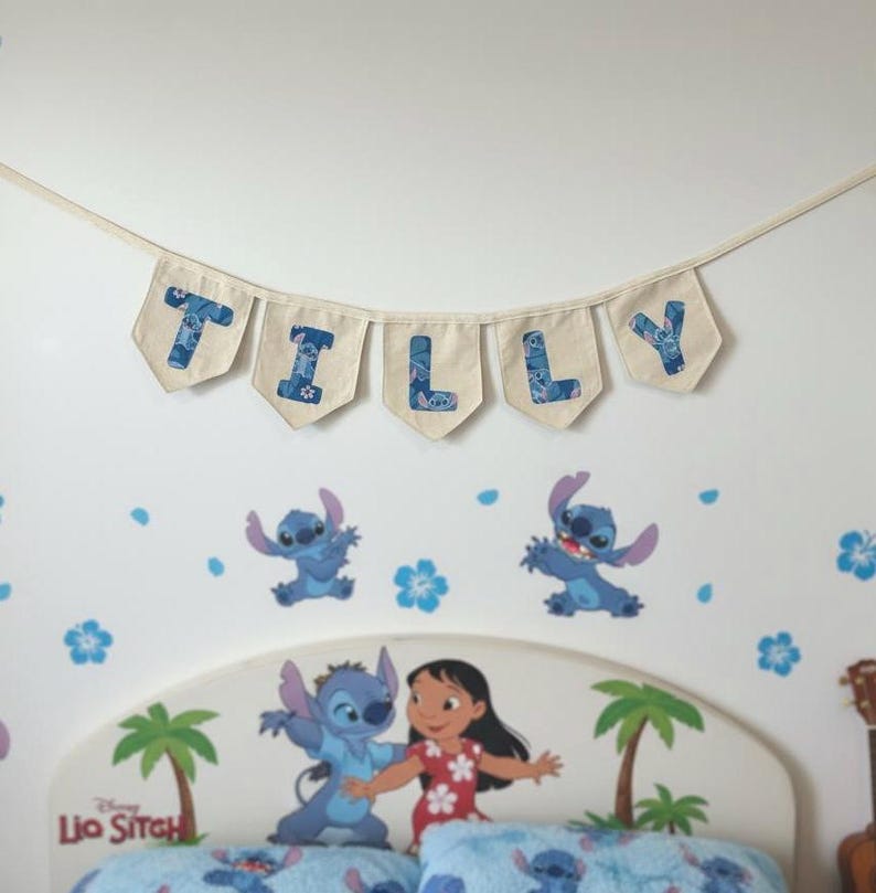 K&ouml;nnte beinhalten: Ein Banner mit der Aufschrift "TILLY" h&auml;ngt &uuml;ber einem Bett mit dem Disney Lilo & Stitch-Thema. Die Buchstaben sind blau auf beigem Hintergrund. Das Kopfteil zeigt Lilo und Stitch, Palmen und die Worte "Lilo & Stitch".