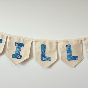 K&ouml;nnte beinhalten: Eine Wimpelkette mit dem Namen "TILLY" in blauen Buchstaben mit einem Stitch-Muster auf einzelnen cremefarbenen Wimpeln. Die Kette ist an einem cremefarbenen Band aufgereiht.