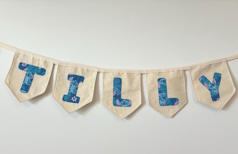K&ouml;nnte beinhalten: Eine Wimpelkette mit dem Namen "TILLY" in blauen Buchstaben mit einem Stitch-Muster auf einzelnen cremefarbenen Wimpeln. Die Kette ist an einem cremefarbenen Band aufgereiht.