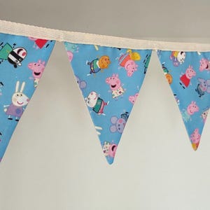 May include: A fabric bunting banner featuring a repeating pattern of Peppa Pig characters on a light blue background. The triangular flags are attached to a cream-coloured ribbon, displaying various cartoon figures in colourful outfits.