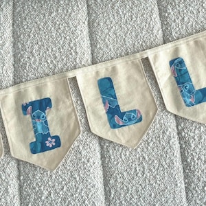 K&ouml;nnte beinhalten: Ein Banner mit der Aufschrift "TILLY" mit cremefarbenen Wimpeln. Jeder Wimpel zeigt einen blauen Buchstaben mit einem Stitch-Charakter-Design. Das Banner ist auf einem wei&szlig;en Band aufgereiht und wird auf einer strukturierten Oberfl&auml;che angezeigt.