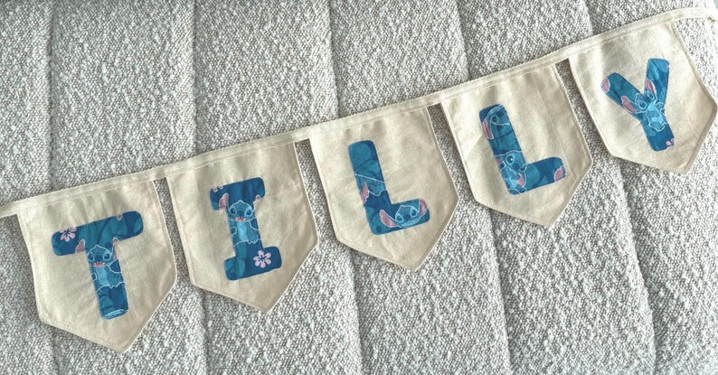 K&ouml;nnte beinhalten: Ein Banner mit der Aufschrift "TILLY" mit cremefarbenen Wimpeln. Jeder Wimpel zeigt einen blauen Buchstaben mit einem Stitch-Charakter-Design. Das Banner ist auf einem wei&szlig;en Band aufgereiht und wird auf einer strukturierten Oberfl&auml;che angezeigt.