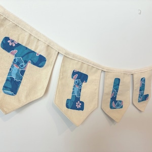 K&ouml;nnte beinhalten: Ein Banner mit der Aufschrift "Tilly" mit einzelnen Flaggen. Jede Flagge ist hellbeige mit einem blau-t&uuml;rkisen Muster mit der Figur Stitch. Die Buchstaben sind ebenfalls blau und t&uuml;rkis.