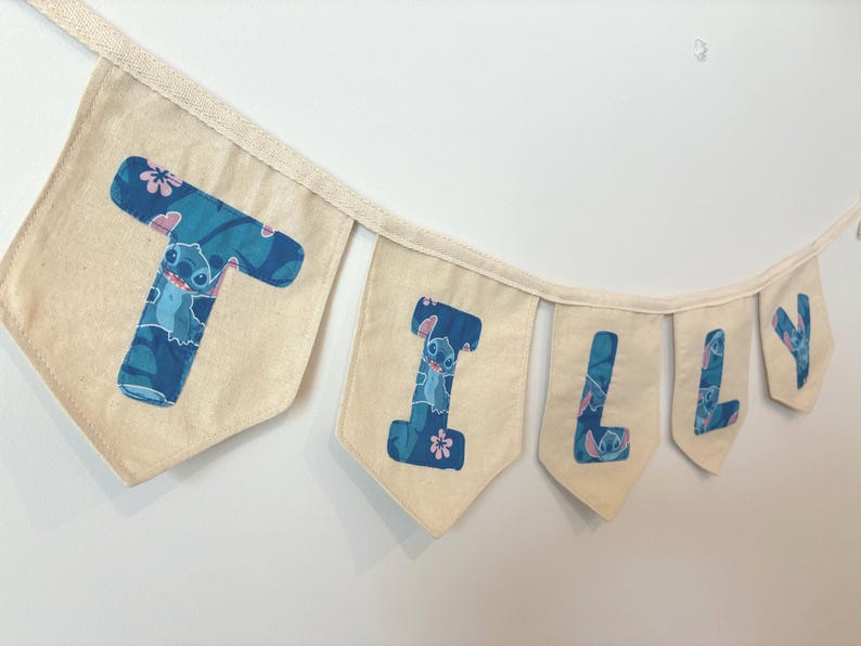K&ouml;nnte beinhalten: Ein Banner mit der Aufschrift "Tilly" mit einzelnen Flaggen. Jede Flagge ist hellbeige mit einem blau-t&uuml;rkisen Muster mit der Figur Stitch. Die Buchstaben sind ebenfalls blau und t&uuml;rkis.