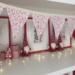 May include: A fabric bunting banner featuring a repeating red line-art pattern of holiday icons including gingerbread men, houses, and candy canes on a white background, suspended from a simple cream-coloured fabric tape string.