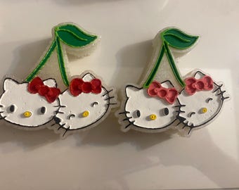 May include: Two Hello Kitty-themed cherry-shaped decorations. Each features two white cat faces with black whiskers, red bows, and yellow eyes. Green leaves and stems complete the design, creating a playful and cute aesthetic.