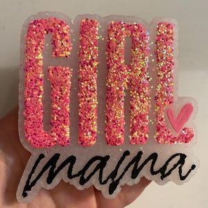 May include: A pink glitter resin piece with the word "GIRL" and "mama" in black script. A pink heart is next to the word "GIRL". The letters are filled with pink glitter. The piece is white around the edges.