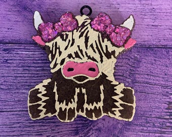 May include: A Highland cow ornament with a black loop for hanging. The cow has a white and brown body, pink nose, and a sparkly pink bow. The background is a purple wooden surface.