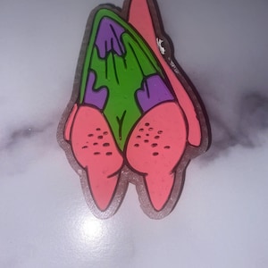 May include: A colorful Patrick Star-shaped charm. The charm features a pink body with a green and purple center. The charm has a clear outline and is likely made of resin or a similar material.