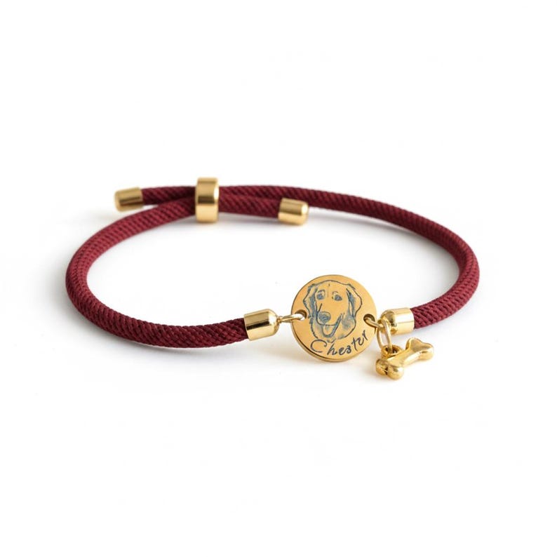 May include: A maroon cord bracelet featuring a circular gold-toned charm engraved with a dog illustration and the name Chester. The bracelet includes gold-toned metal end caps, a sliding adjustment bead, and a small gold-toned bone-shaped charm.
