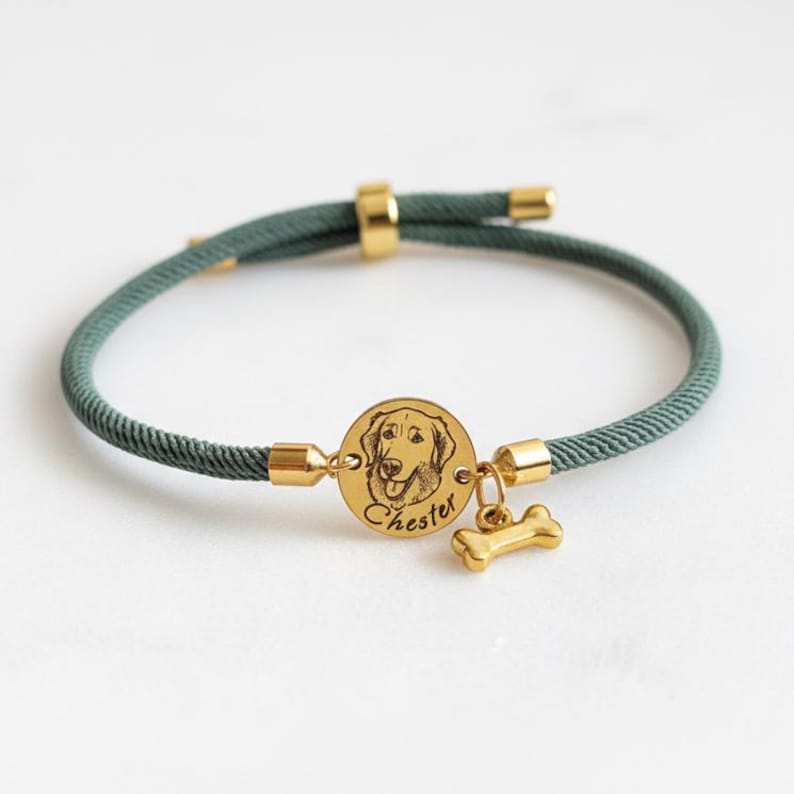 May include: A dark green cord bracelet featuring a circular gold-toned metal charm engraved with a dog portrait and the name Chester, accompanied by a small gold-toned bone-shaped charm, with gold-toned metal end caps and an adjustable sliding clasp.