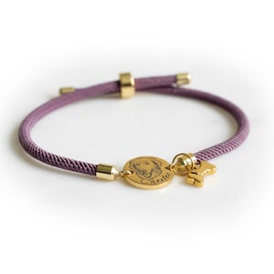May include: A purple cord bracelet featuring a gold-toned circular charm engraved with a dog illustration and the name Chester, accompanied by a small gold-toned bone-shaped charm. The bracelet includes gold-toned metal end caps and a sliding clasp.