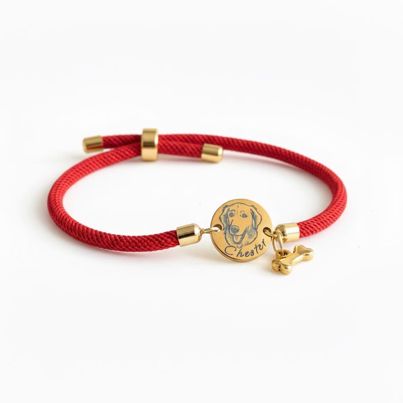 May include: A red cord bracelet featuring a circular gold-toned metal charm engraved with a dog illustration and the name Chester, accompanied by a small gold-toned bone-shaped charm and adjustable gold-toned metal end caps and sliding bead.