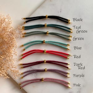 May include: A collection of eight cord bracelets in black, teal, green, blue, red, dark red, purple, and pink. Each bracelet features two parallel cords joined by a central gold-toned metal bead and finished with gold-toned metal end caps.