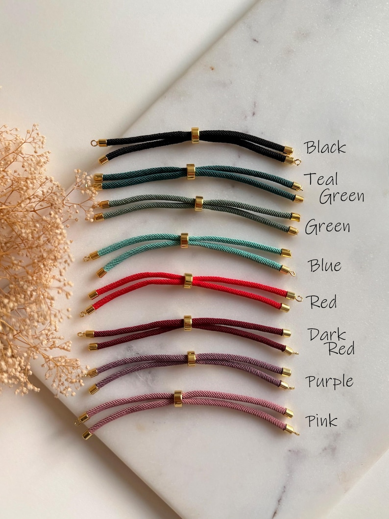 May include: A collection of eight cord bracelets in black, teal, green, blue, red, dark red, purple, and pink. Each bracelet features two parallel cords joined by a central gold-toned metal bead and finished with gold-toned metal end caps.