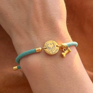 May include: A teal cord bracelet featuring a circular gold-toned charm engraved with a dog portrait and the name Chester, accompanied by a small gold-toned bone-shaped charm, finished with gold-toned metal end caps and adjustable hardware.