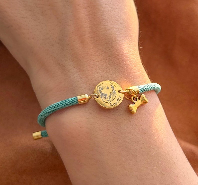 May include: A teal cord bracelet featuring a circular gold-toned charm engraved with a dog portrait and the name Chester, accompanied by a small gold-toned bone-shaped charm, finished with gold-toned metal end caps and adjustable hardware.