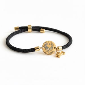 May include: A black cord bracelet featuring a circular gold-toned metal charm engraved with a dog portrait and the name Chester, accompanied by a small gold-toned bone-shaped charm, with gold-toned metal accents and an adjustable sliding closure.