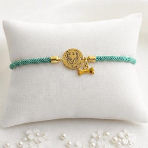 May include: A teal cord bracelet featuring a circular gold-toned charm engraved with a dog portrait and the name Chester, accompanied by a small gold-toned bone-shaped charm. The bracelet is secured with gold-toned metal end caps on each side.