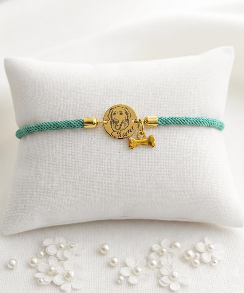 May include: A teal cord bracelet featuring a circular gold-toned charm engraved with a dog portrait and the name Chester, accompanied by a small gold-toned bone-shaped charm. The bracelet is secured with gold-toned metal end caps on each side.