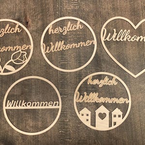 Puede incluir: Cinco letreros de bienvenida de madera con diferentes diseños. Los letreros son todos redondos excepto uno que tiene forma de corazón. Los letreros dicen "Herzlich Willkommen" en alemán.