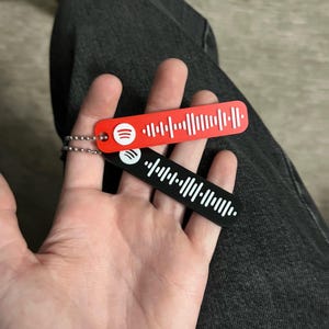 Könnte beinhalten: Zwei Spotify-Code-Schlüsselanhänger, einer rot und einer schwarz, werden in einer Hand gehalten. Jeder Schlüsselanhänger hat ein weißes Spotify-Logo und ein weißes, strichcodeähnliches Design. Die Schlüsselanhänger sind an kleinen Silberketten befestigt.