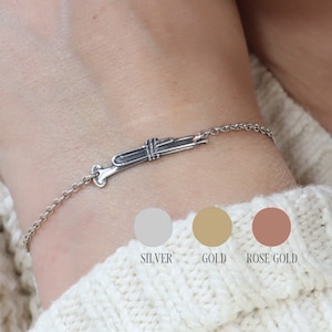 May include: A silver bracelet with a trumpet charm. The bracelet is shown on a person's wrist. The image also shows three colour swatches: silver, gold, and rose gold.
