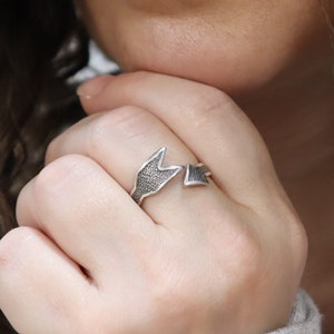 May include: A silver ring with a unique design featuring two interlocking arrowheads. The ring is worn on a person's finger.