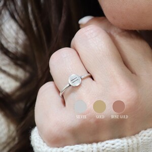 May include: A silver ring with a thin band and a round, flat top featuring two parallel lines. The ring is shown on a person's finger. The image also includes color swatches for silver, gold, and rose gold.