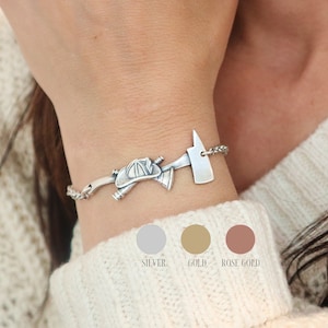 May include: A silver bracelet featuring a firefighter's helmet and axe charm. The bracelet is on a delicate chain and is displayed on a person's wrist. The image also shows color options: silver, gold, and rose gold.