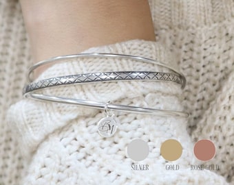 May include: Three silver bangle bracelets with a textured design and a small charm with the initials "BW" hanging from the bottom. The bracelets are shown on a person's wrist. The image also shows three color swatches for the bracelets: silver, gold, and rose gold.