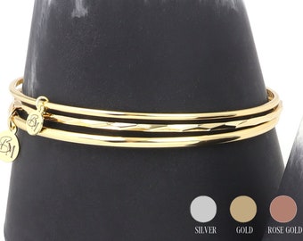 Könnte beinhalten: Drei goldene Armreifen mit einem kleinen Anhänger. Der Anhänger ist rund und hat den Buchstaben "B" eingraviert. Die Armreifen sind übereinander gestapelt und auf einem schwarzen, kegelförmigen Ständer ausgestellt. Der Ständer steht auf einer weißen Oberfläche. Das Bild zeigt auch drei Farbmuster: Silber, Gold und Roségold.