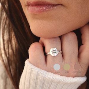 May include: A silver ring with a round, flat top featuring two parallel lines. The ring is shown on a person's finger. The image also shows three color swatches: silver, gold, and rose gold.